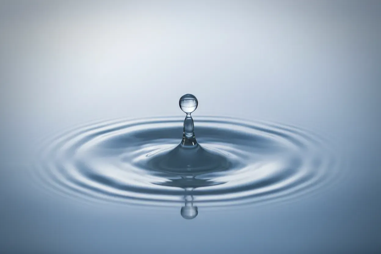 Macro of a single water droplet creating concentric ripples on calm water, set against a soft blue-gray gradient background.